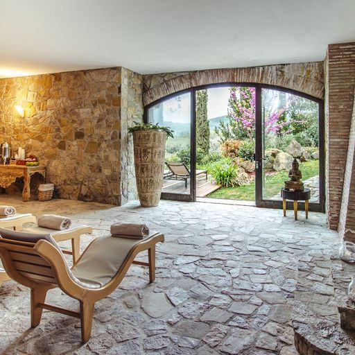 Rustic spa room with stone walls, lounge chairs, and a large window overlooking a lush garden. Soft lighting creates a tranquil atmosphere.