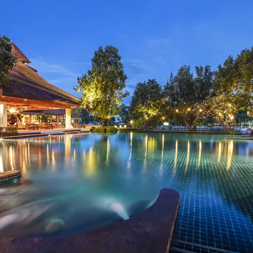 A serene resort pool at dusk, surrounded by lush trees and illuminated by soft lights, with a thatched-roof building and lounge chairs nearby.