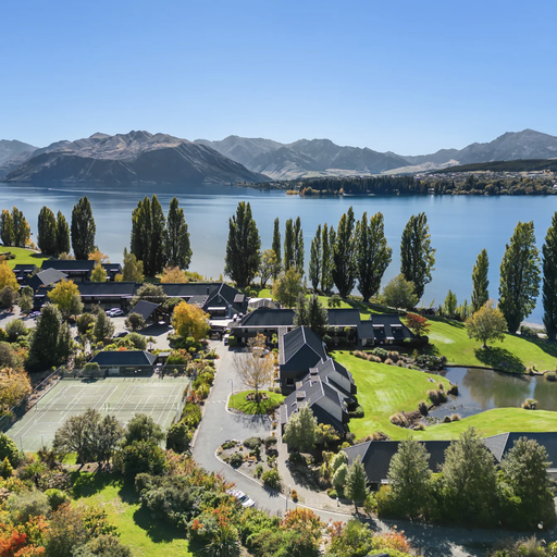 Aerial view of a lakeside resort with lush greenery, tennis court, and mountains in the background under a clear blue sky.
