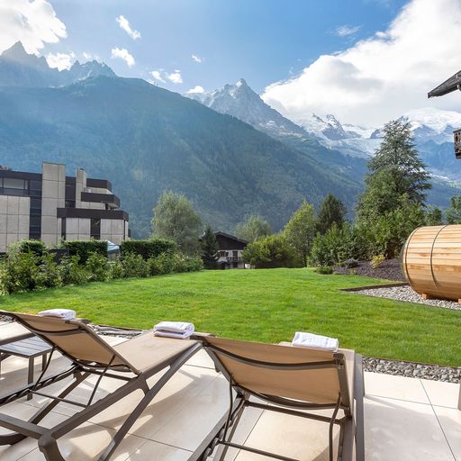 Lounge chairs and a sauna overlook a grassy area with mountain views under a partly cloudy sky.