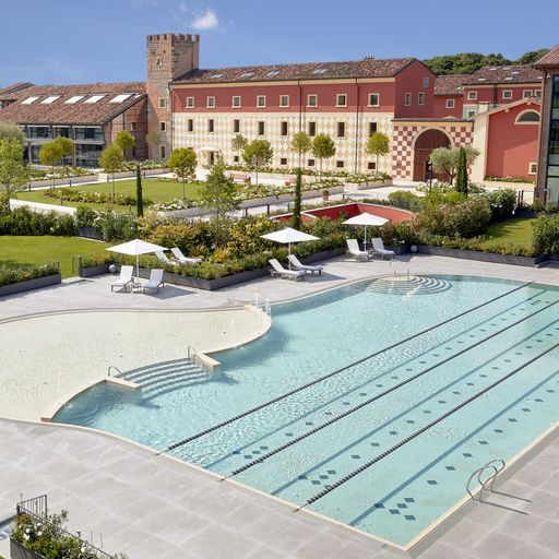 Outdoor swimming pool with lanes, surrounded by greenery and lounge chairs. Historic building with red and beige facade in the background.