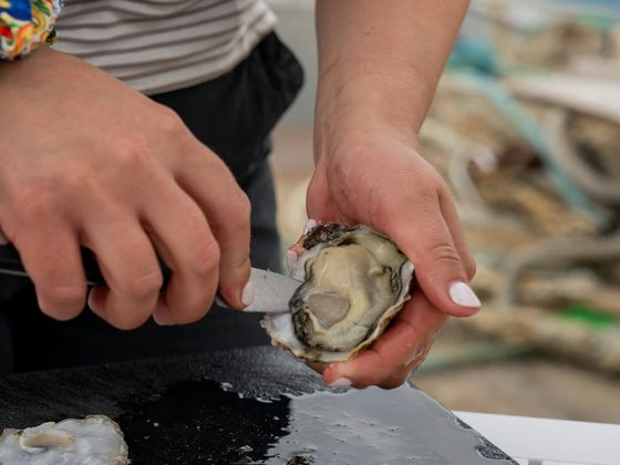 A person in a striped shirt shucks an oyster on a black cutting board, with one oyster half already opened and placed nearby.