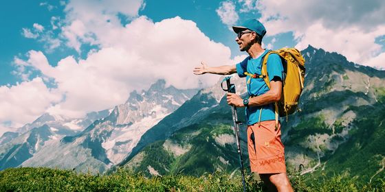 A hiker with a yellow backpack gestures towards a mountain range under a blue sky with clouds.