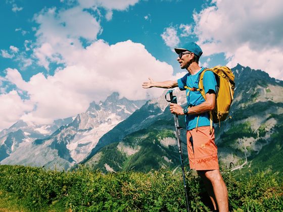 A hiker with a yellow backpack gestures towards a mountain range under a partly cloudy sky, standing on a lush green hillside.