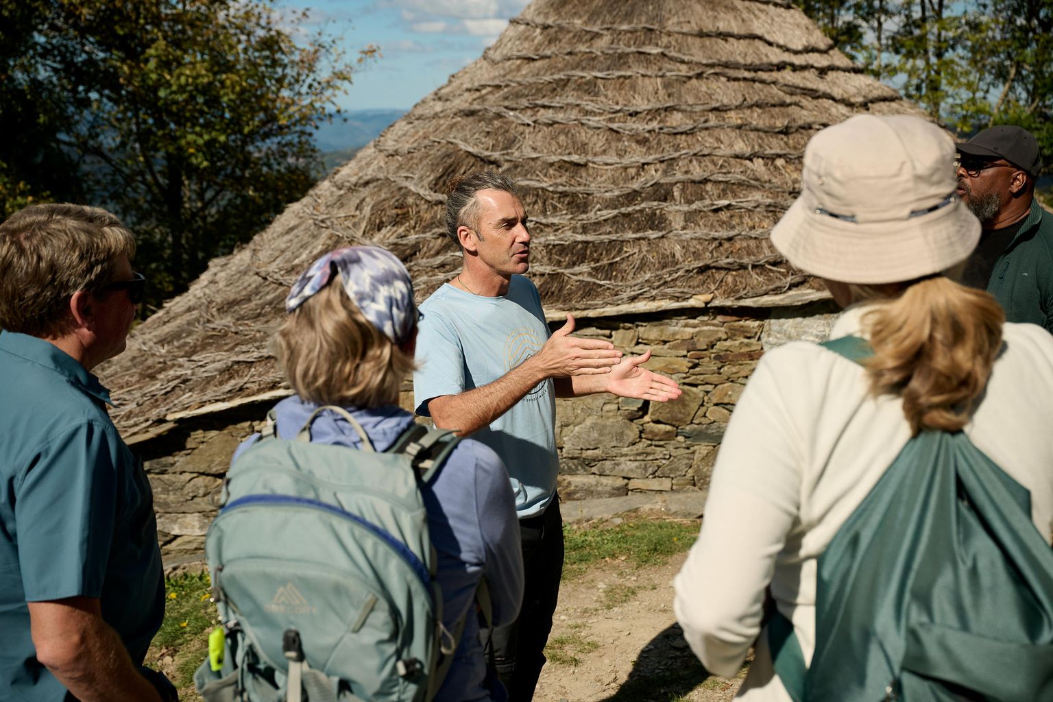 A guide talks to a group of hikers near a stone and thatch structure, with trees and a blue sky in the background.