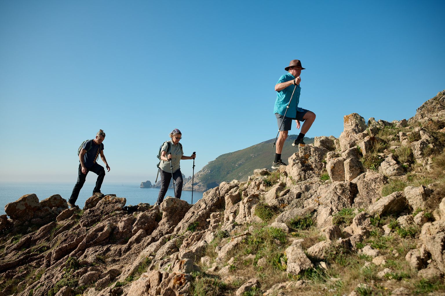 Three hikers with trekking poles ascend a rocky hill, with a scenic ocean view in the background under a clear blue sky.