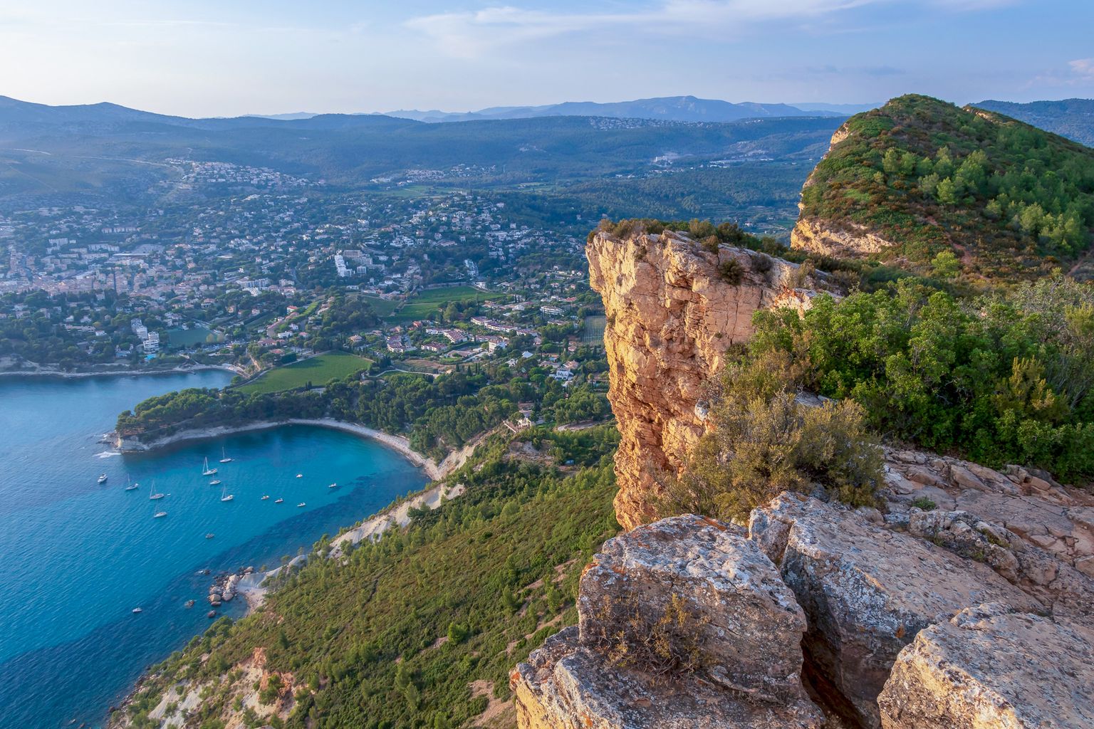 Aerial view of a rugged cliff overlooking a coastal town with a turquoise bay, boats, and lush greenery under a clear sky.