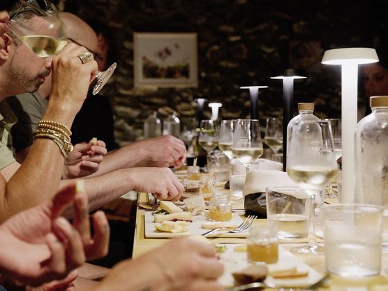 People enjoying a meal at a dimly lit table, with wine glasses and plates of food, illuminated by modern table lamps.