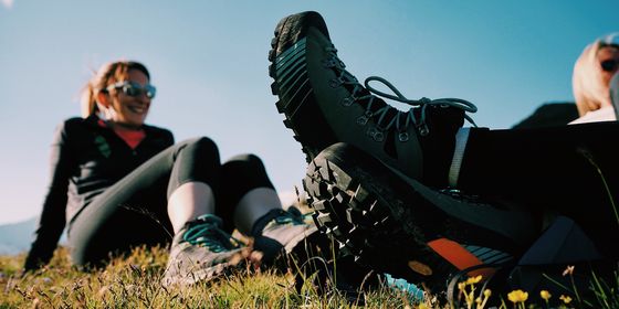 Two people sitting on grass in hiking gear, with focus on their boots. Bright blue sky and sunlight create a relaxed outdoor atmosphere.