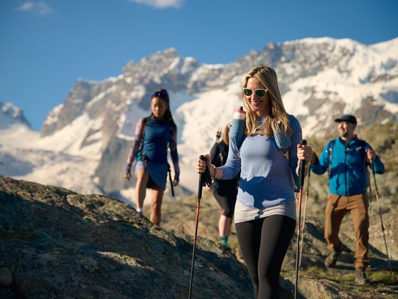 A group of hikers with trekking poles walk on rocky terrain, with snow-capped mountains in the background under a clear blue sky.