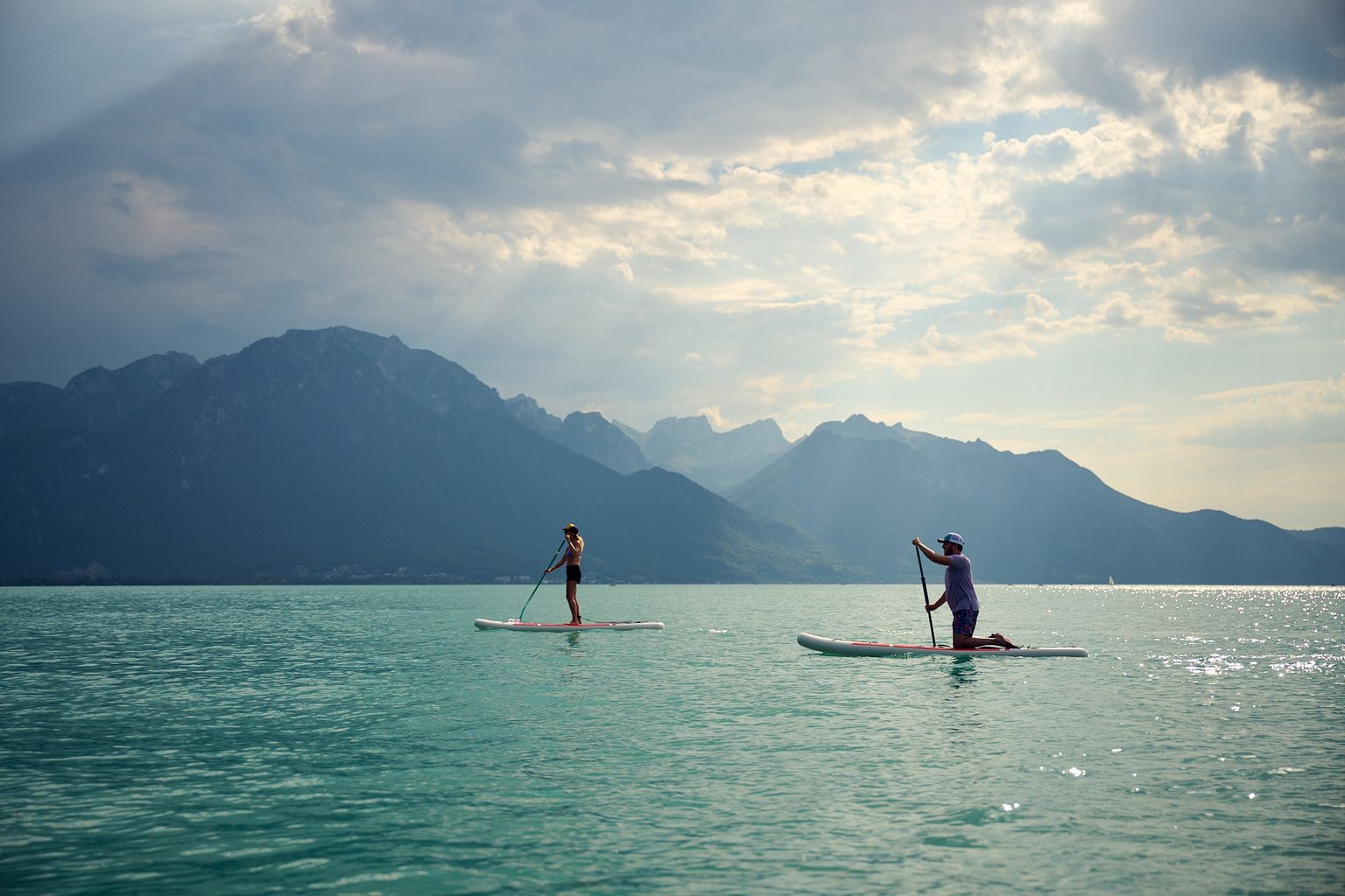 Two people paddleboarding on a calm lake with mountains in the background under a cloudy sky.