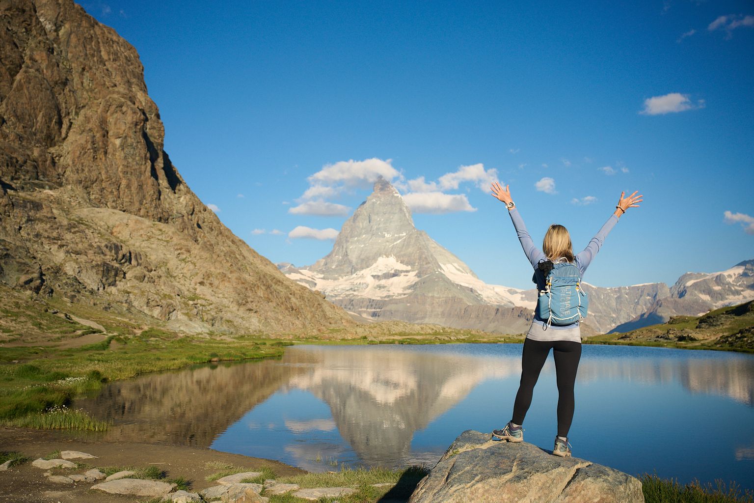 A person with a backpack stands on a rock, arms raised, facing a mountain reflected in a clear lake under a blue sky with scattered clouds.