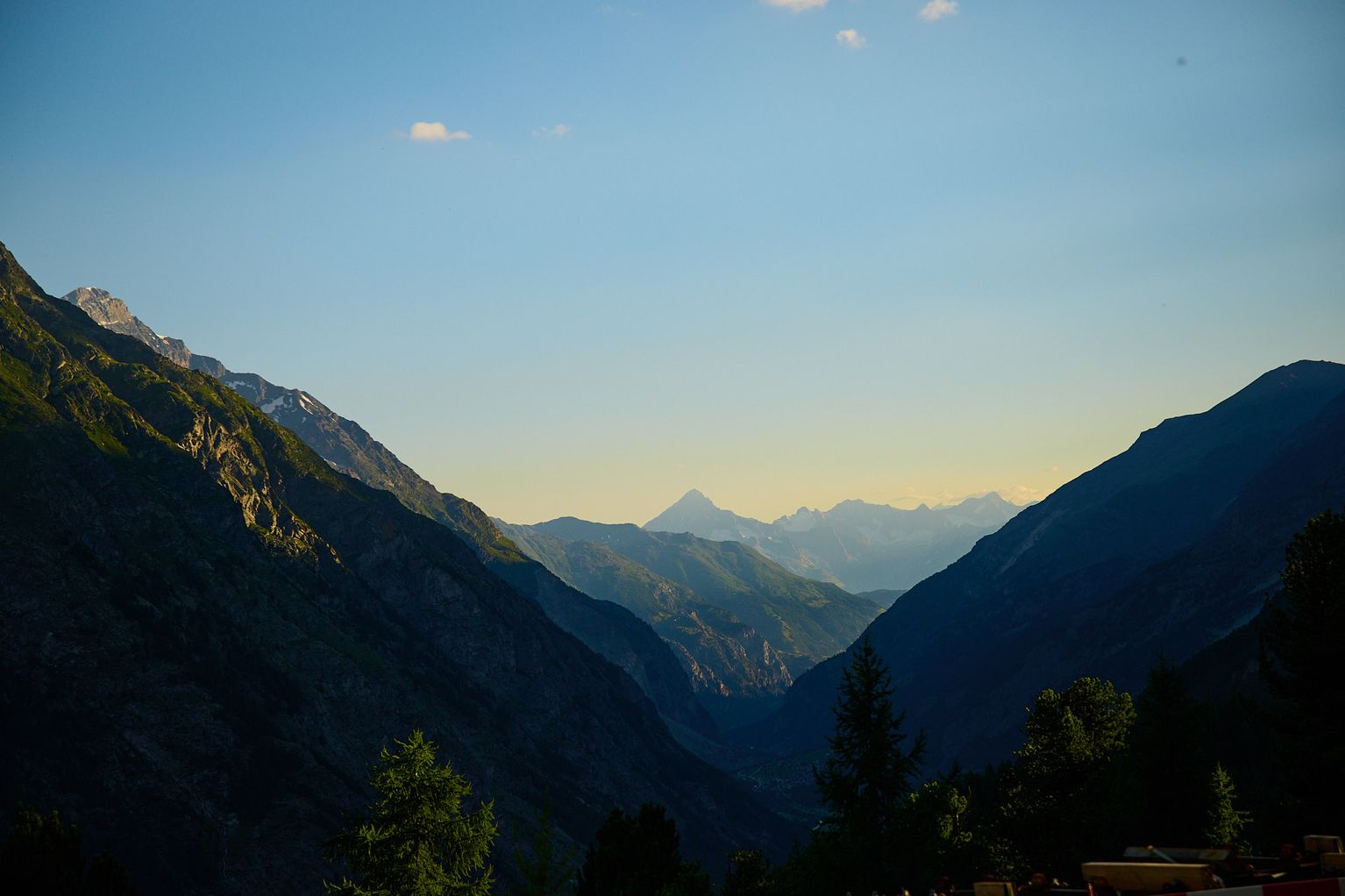 Mountain landscape at sunset with a view of distant peaks and a clear sky, framed by darkened valleys and scattered trees in the foreground.