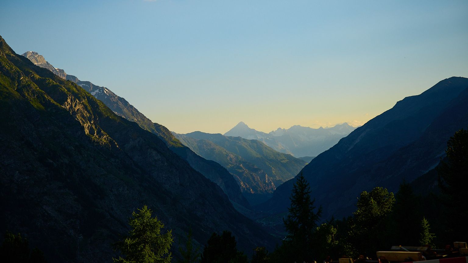 Mountain landscape at sunset with a view of distant peaks and a clear sky, framed by darkened valleys and scattered trees in the foreground.