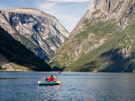 Two people kayak on a calm fjord, surrounded by towering, rocky cliffs under a clear sky.