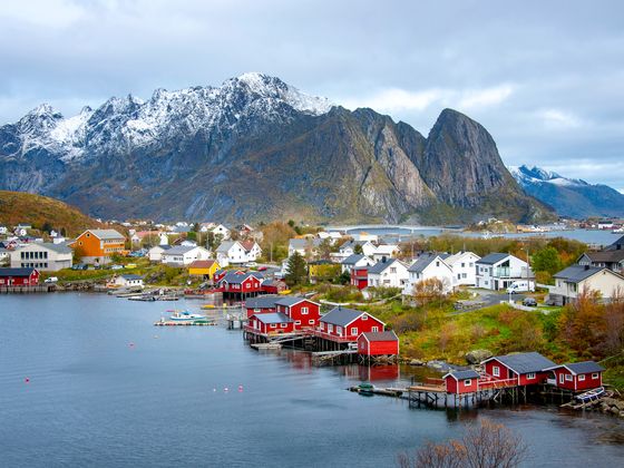 Scenic view of a coastal village with red and white houses near a calm bay, set against snowy mountains under a cloudy sky.