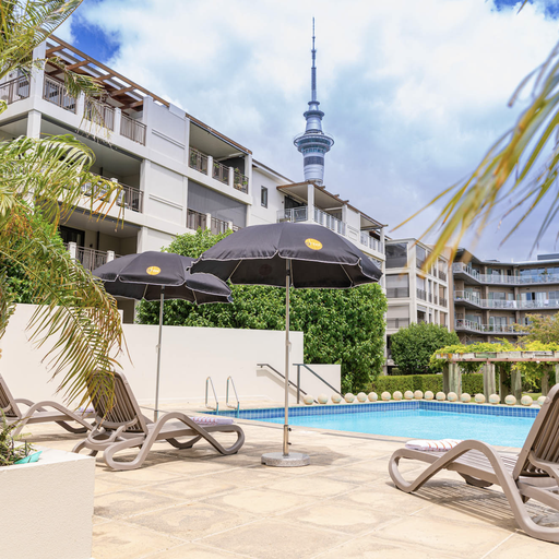 Poolside scene with lounge chairs, umbrellas, and palm trees, set against modern buildings and a tall tower under a partly cloudy sky.