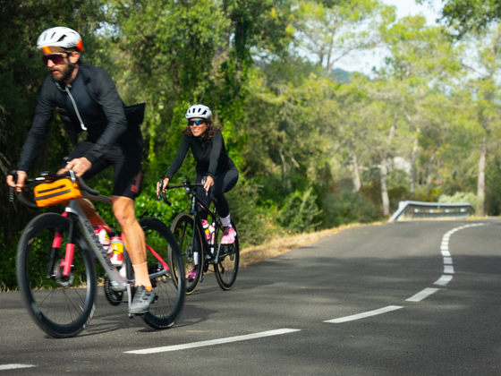 Two cyclists wearing helmets ride on a sunny, tree-lined road, with one ahead of the other.