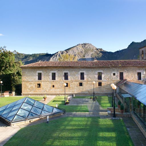 Historic stone building with a courtyard, glass pyramid structure, and mountainous backdrop under a clear blue sky.