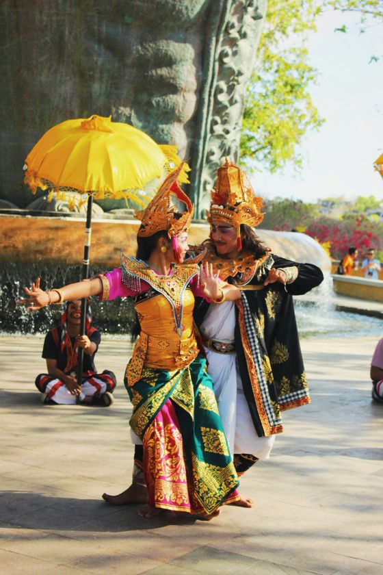 Two dancers in traditional Balinese attire perform gracefully outdoors, with a large statue and yellow umbrella in the background.