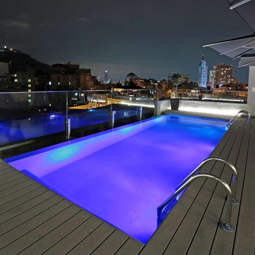 Rooftop pool with glowing blue water, surrounded by a wooden deck and umbrellas, overlooking a city skyline at night.