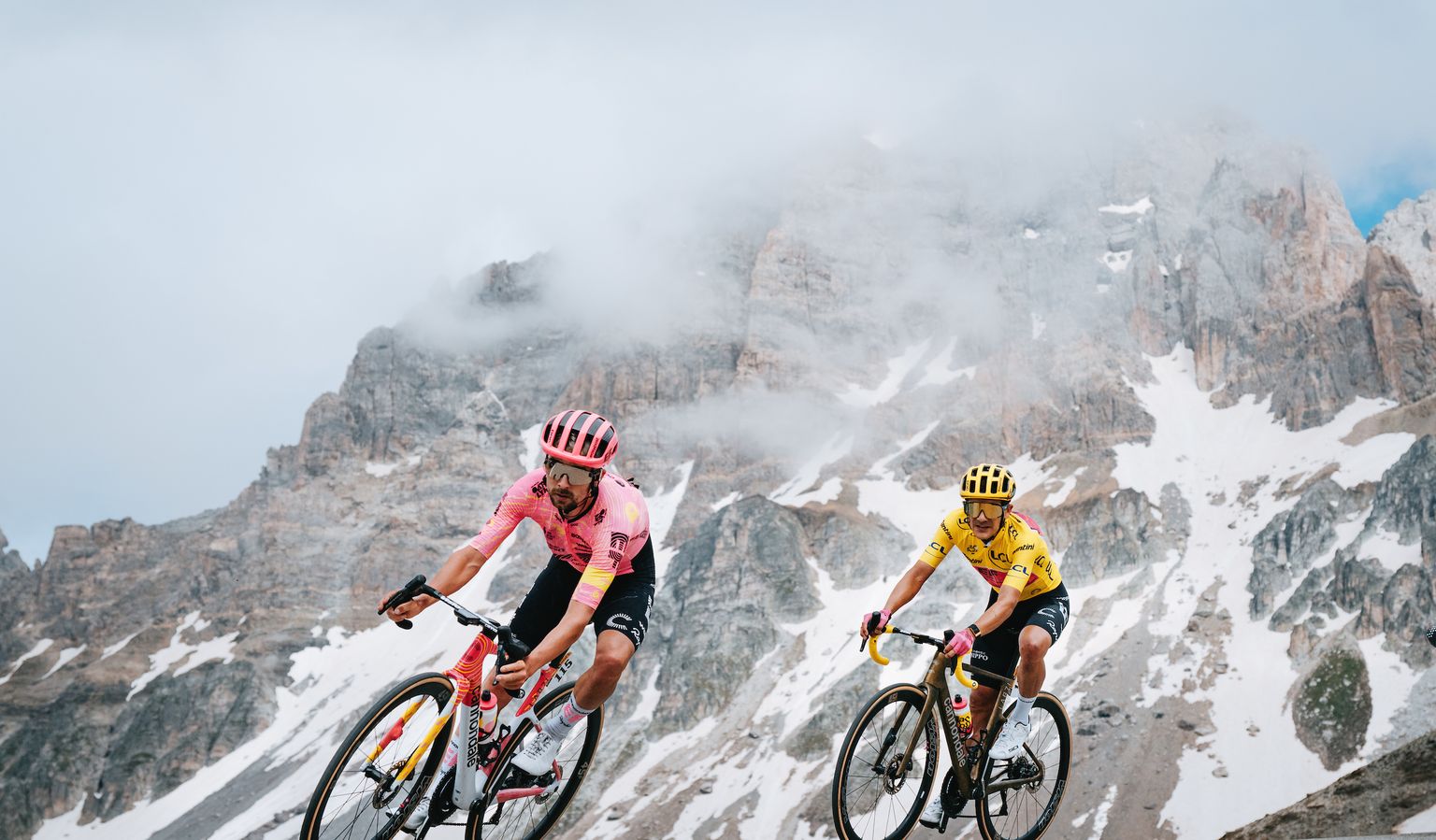 Two cyclists in colorful jerseys race through a mountainous landscape under a cloudy sky, with snow patches visible on the rugged peaks.