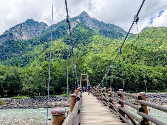 Wooden suspension bridge leading to a lush green forest with a towering mountain in the background under a cloudy sky.