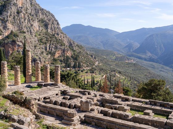 Ancient ruins with stone columns at Delphi, Greece, set against a backdrop of rugged mountains and lush valley under a clear blue sky.