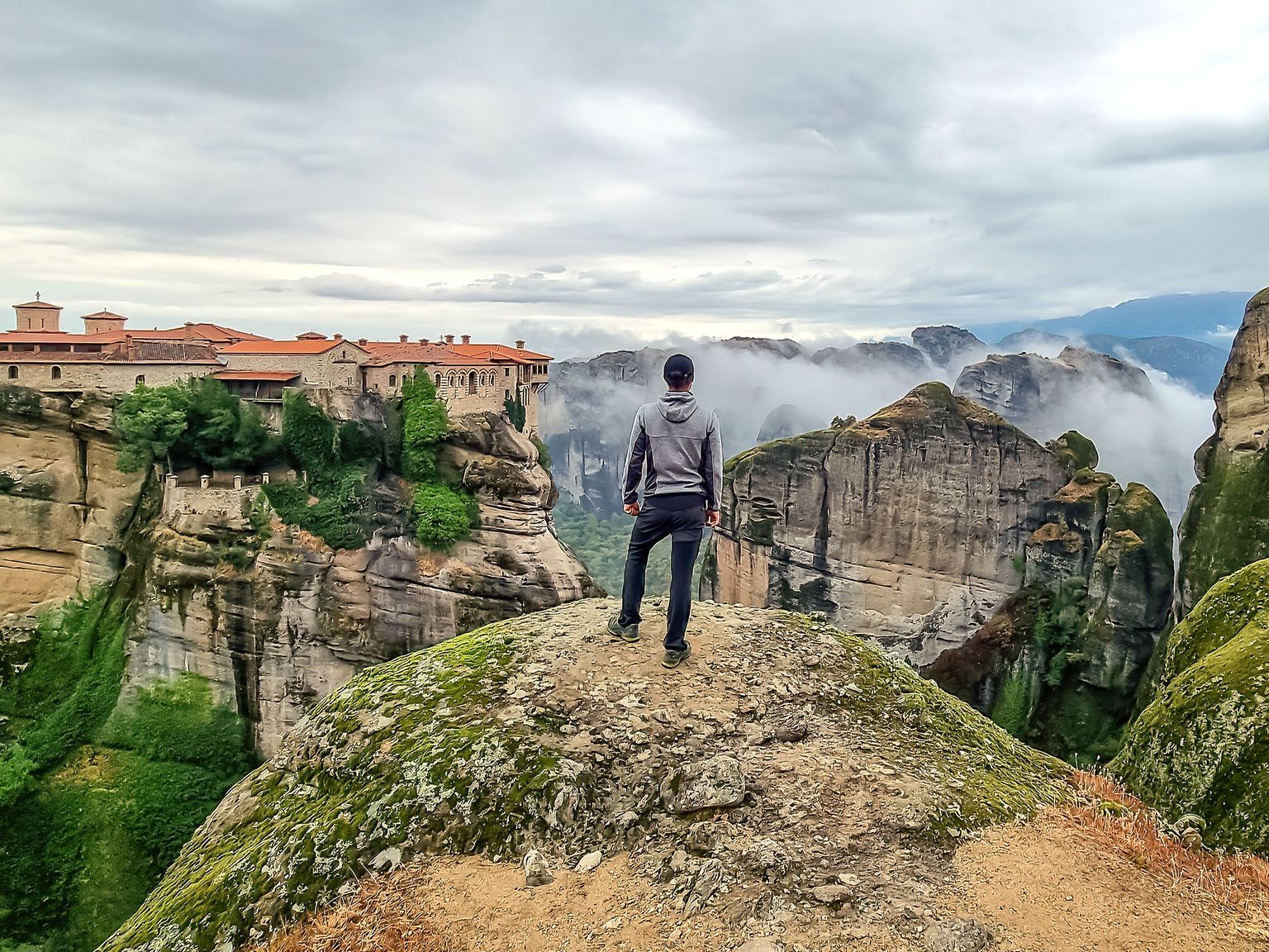 A person stands on a cliff overlooking monasteries built atop towering rock formations surrounded by misty valleys and cloudy skies.