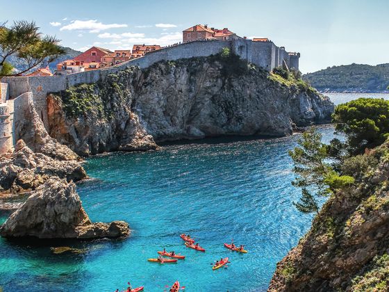 Kayakers paddle in clear blue water near scenic cliffs and historic fortified walls, with a Mediterranean landscape in the background.