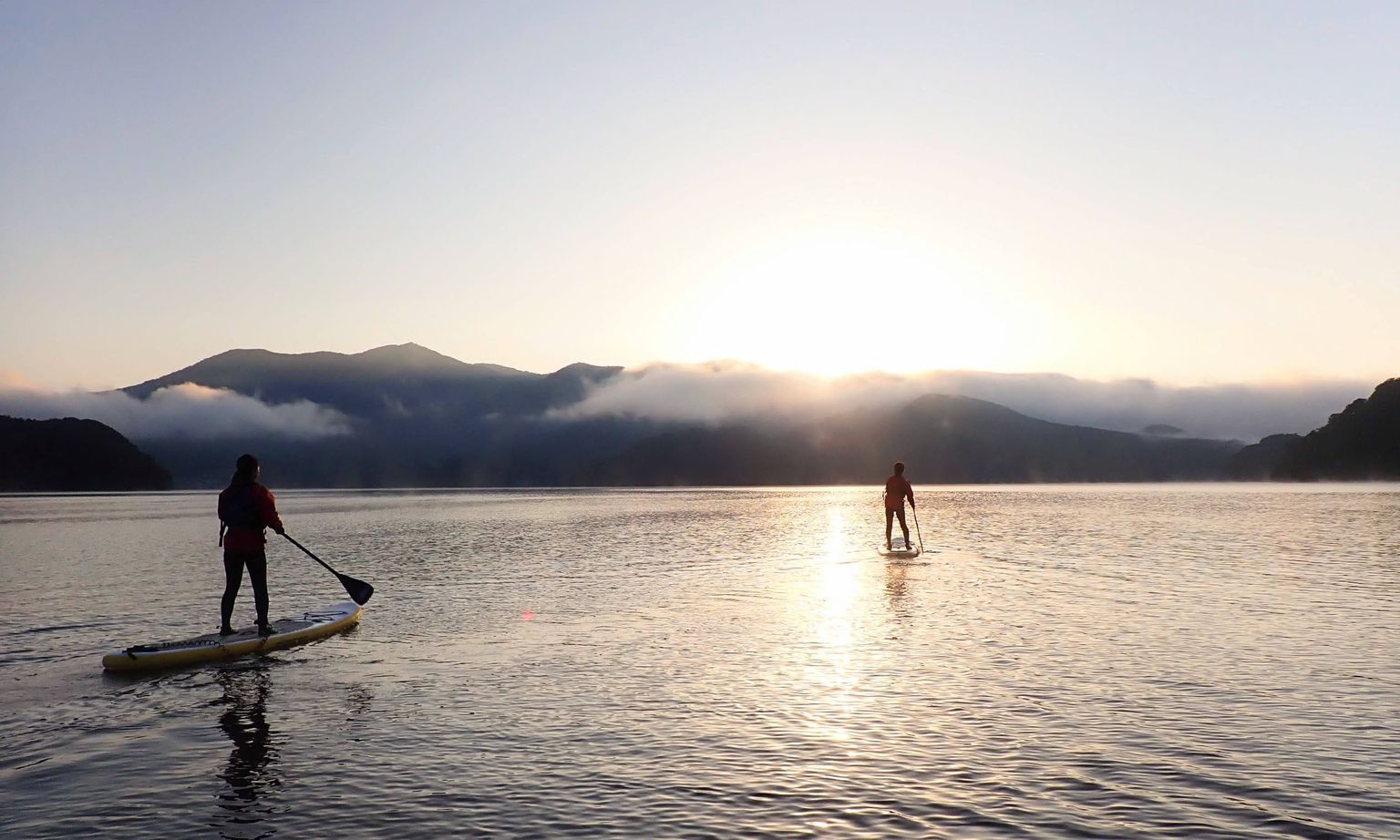 Two people paddleboarding on a calm lake at sunrise, surrounded by mist-covered mountains in the background.