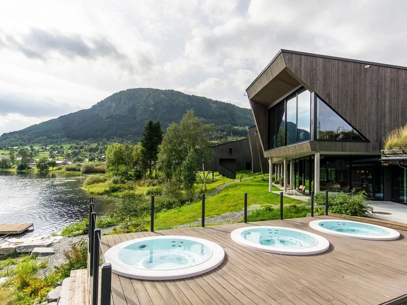Modern building with large windows, three outdoor hot tubs on a wooden deck, and a scenic view of a lake and mountain under a cloudy sky.