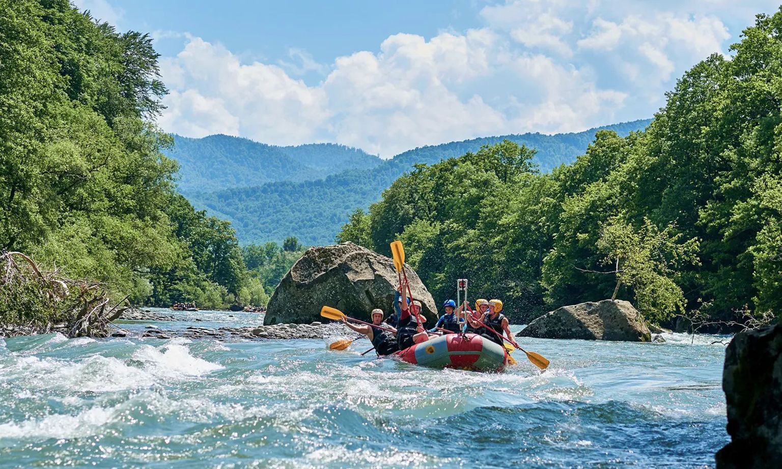 Group of people white-water rafting through a river surrounded by lush green trees and distant mountains under a blue sky with clouds.