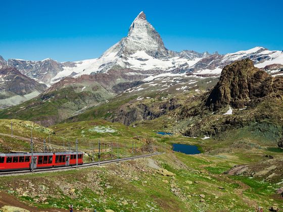 A red train travels through green hills with the snow-capped Matterhorn in the background under a clear blue sky.