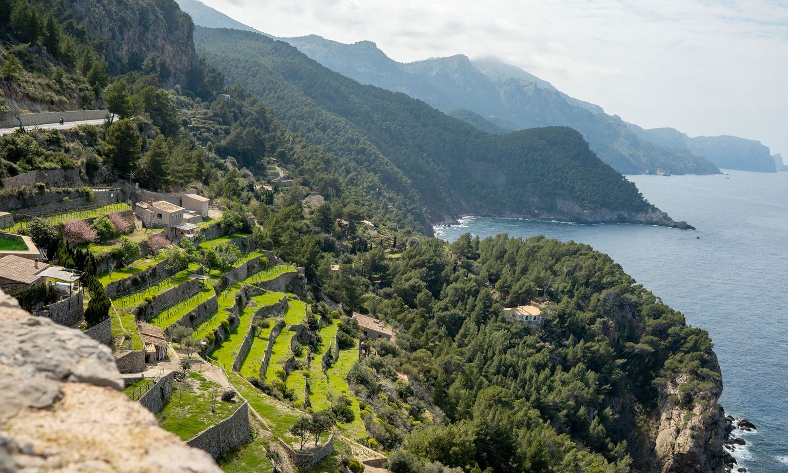 Terraced green fields and rustic houses on a coastal hillside, with cliffs and a calm sea under a partly cloudy sky.