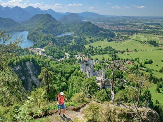 Hiker with a red backpack stands on a cliff overlooking Neuschwanstein Castle, surrounded by lush greenery and mountains under a blue sky.