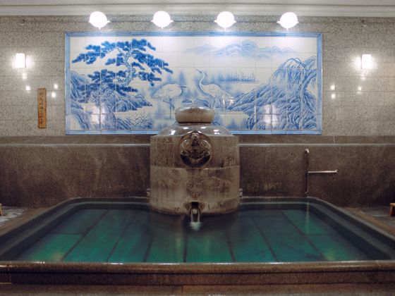 Indoor public bath with a tile mural depicting cranes and mountains, featuring a central stone fountain and surrounded by faucets with stools.