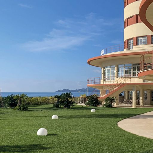 Curved modern building with balconies by a lush green lawn, overlooking the sea under a clear blue sky.