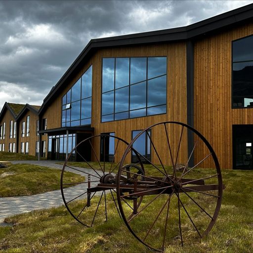 Modern wooden building with large windows under a cloudy sky. An old metal wagon wheel sculpture sits on a grassy area near a stone path.