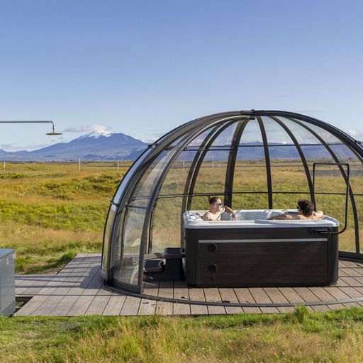 Two people relax in a hot tub under a clear dome on a wooden deck, surrounded by open grassy fields and distant mountains under a blue sky.