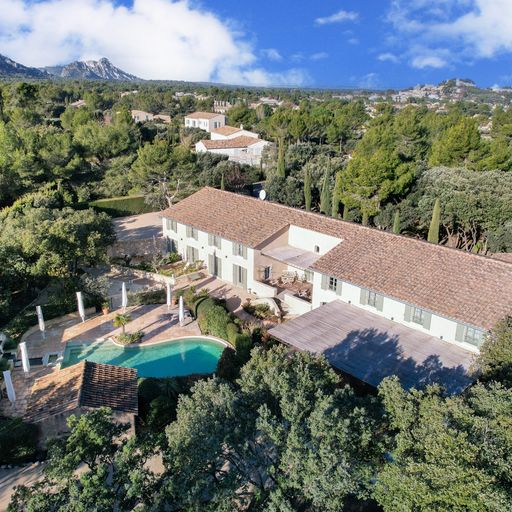 Aerial view of a large villa with a terracotta roof, surrounded by trees, featuring a pool and patio, set against a mountainous backdrop.