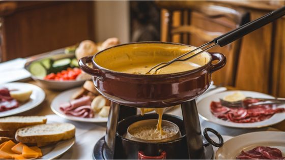 A pot of cheese fondue with a fork, surrounded by plates of bread, meats, and vegetables on a dining table.