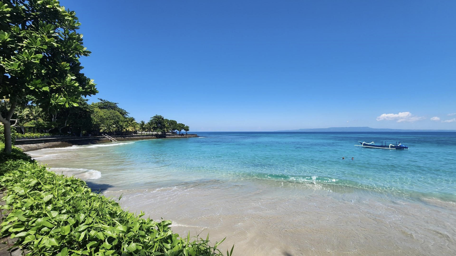 A serene beach with turquoise water, a small boat, and lush greenery under a clear blue sky. Two people swim near the shore.