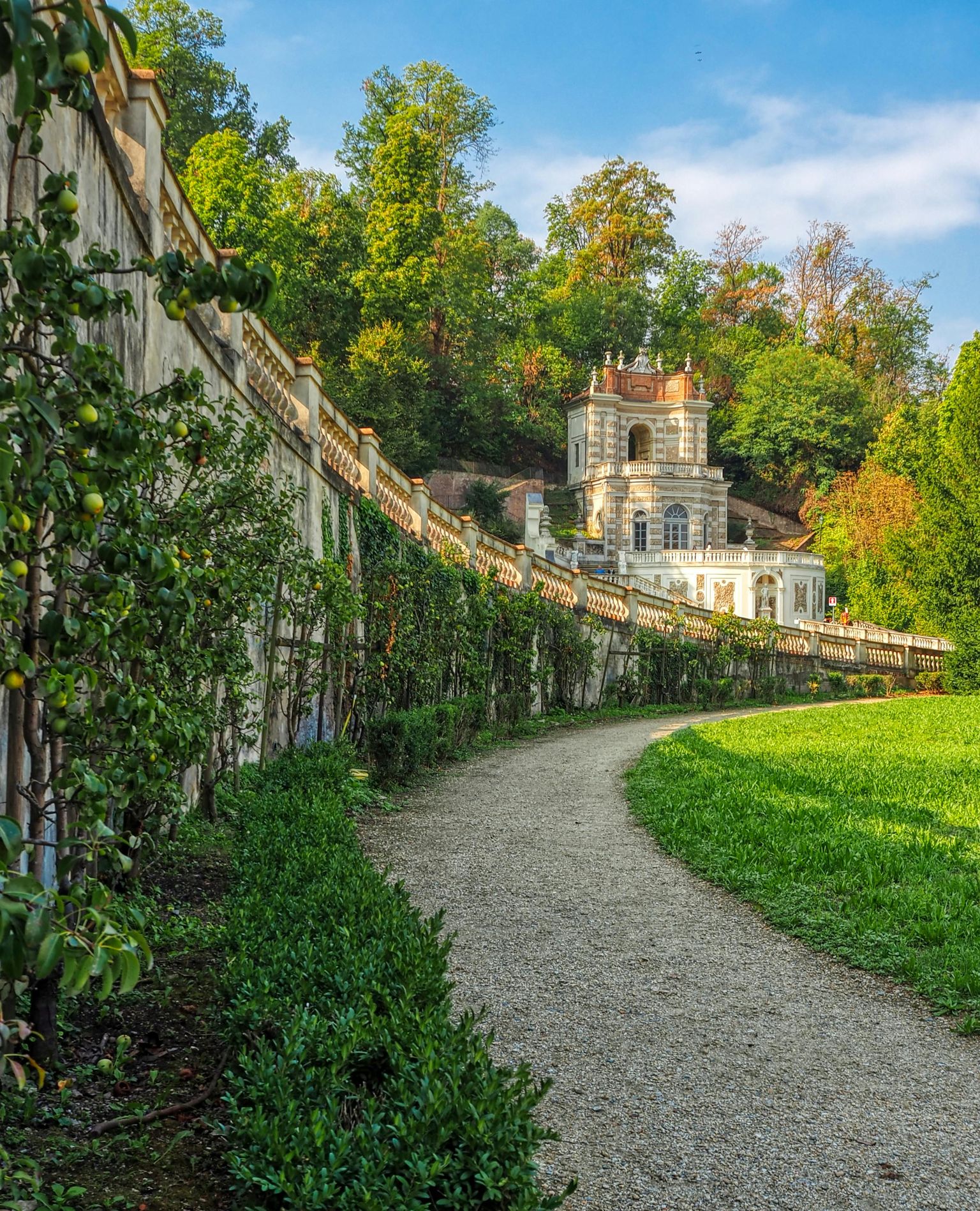 A gravel path leads to an ornate villa surrounded by lush greenery and a stone wall, set against a backdrop of trees under a clear blue sky.
