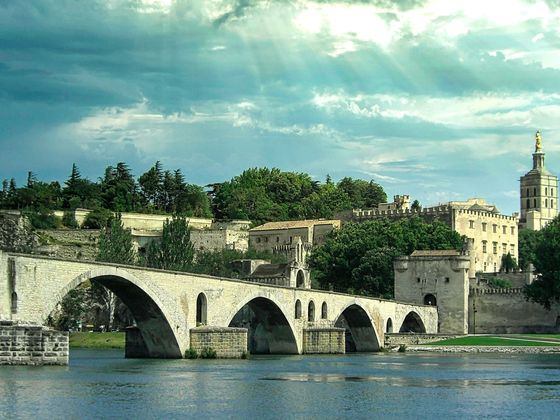 Historic stone bridge over a river with arches, leading to an ancient fortified city with towers and lush greenery under a cloudy sky.