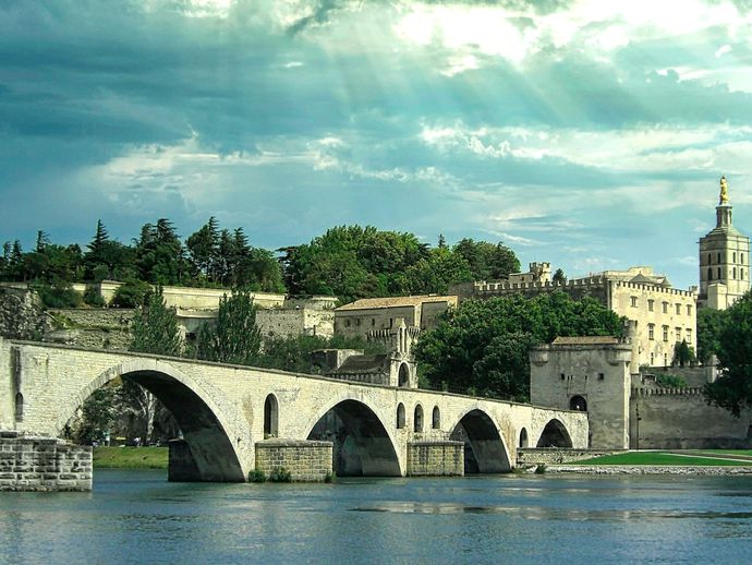 Historic stone bridge over a river with arches, leading to an ancient fortified city with towers and lush greenery under a cloudy sky.