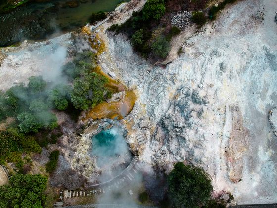 Aerial view of a geothermal area with steaming hot springs, mineral deposits, and surrounding greenery.