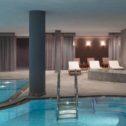 Indoor spa with a modern pool, metal ladder, loungers, and soft lighting. Gray curtains and walls create a serene ambiance.