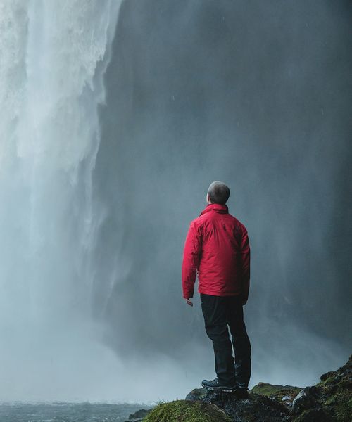 A person in a red jacket stands on a mossy rock, gazing at a powerful waterfall with mist rising around them.
