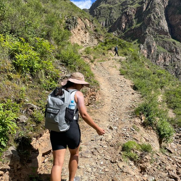 A person wearing a hat and backpack hikes up a rocky mountain trail surrounded by greenery, with another hiker visible ahead.